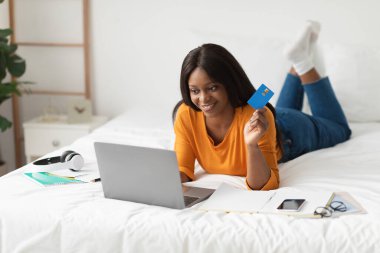 Bir kadın yatağında uzanmış, bir elinde kredi kartı, diğerinde dizüstü bilgisayar tutarken gülümsüyor. Çevrimiçi alışverişle meşgul ve rahat görünüyor..
