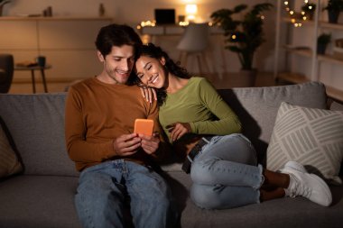 Genç bir çift oturma odalarındaki bir kanepede rahatça oturup bir anı paylaşıyorlar. Mutlu bir şekilde akıllı telefona bakıyorlar, rahatlatıcı bir hafta sonu akşamının tadını çıkarıyorlar..