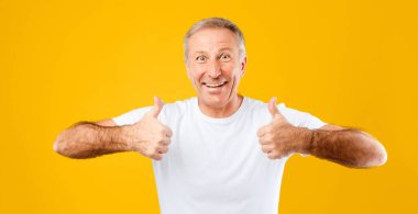 Bir adam parlak sarı bir arka plana karşı duruyor, geniş bir gülümseme ve iki başparmak işareti veriyor. İfadesinde ve duruşunda coşku ve mutluluk gösteriyor..