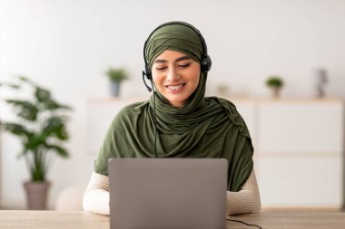 Başörtülü genç Arap kadın evde dizüstü bilgisayarıyla çalışırken gülümsüyor. Kulaklık takıyor, bağımsız olarak çalışıyor, modern uzak kariyerini sergiliyor..