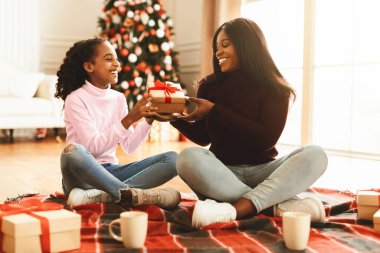 Bir anne ve kızı oturma odasındaki halıya oturmuş, birbirlerine hediye veriyorlar. Odada ışıkları ve süslemeleri olan bir Noel ağacı var. O an için mutlu ve heyecanlı görünüyorlar..