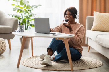 Dizüstü bilgisayarı ve kulaklığı ile yerde oturan bir kadın, mutlu bir şekilde sanal bir toplantıyla meşgul. Etrafı bitkilerle çevrili ve rahat bir oturma odası var..