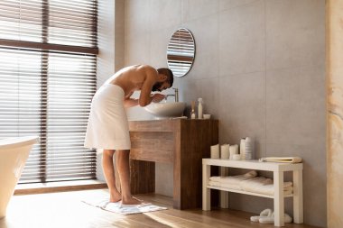 Orta Doğulu kaslı bir adam modern bir banyoda lavabonun yanında duruyor, yüzünü yıkamak için su kullanıyor. Gün ışığının pencereden içeri akması için hazırlanıyor ve yeni bir sabah atmosferi yaratıyor..