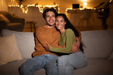 Bir çift rahat bir kanepede otururken gülümsüyor ve birbirlerine sarılıyorlar. Odadan gelen sıcak ışık rahat bir atmosfer yaratıyor. Akşamları neşelerini ve bağlantılarını vurguluyor..