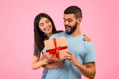 Genç bir çift neşeyi paylaşırken içlerinden biri güzel bir hediyeyi kırmızı kurdeleyle tutuyor. Gülümsüyorlar ve pembe bir arka planla çevrili mutlu bir anın tadını çıkarıyorlar..