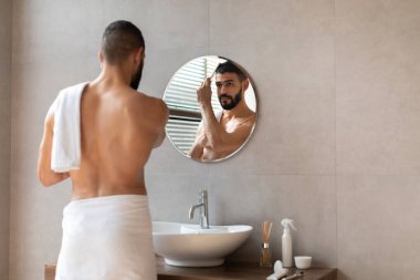 Genç bir adam şık bir banyoda durmuş, yuvarlak aynaya bakıyor. O, beyaz bir havluya sarılı iken ahşap bir tarakla saçını tarıyor. Güneş ışığı odanın içinden süzülüyor.