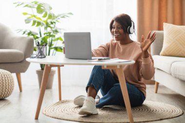 Bir kadın oturma odasında yerde oturuyor, kulaklık takıyor ve bilgisayarında video sohbeti yaparken gülümsüyor. Parlak güneş ışığı boşluğu doldurur ve sıcak bir atmosfer yaratır..