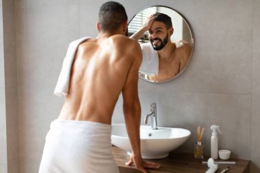 Genç adam duştan sonra saçına dokunurken aynadaki yansımasına gülümsüyor. Üstsüz ve şık bir banyoda duruyor. Bakım rutinine odaklanıyor..