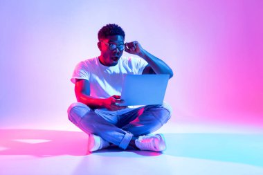 Şok olmuş bir siyah adam dizüstü bilgisayarla yerde oturuyor, neon ışıklı efektlerle çevrili online piyangoyu kazandıktan sonra hayretle gözlüklerine dokunuyor..