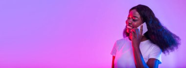 Genç bir Afrikalı Amerikalı kadın akıllı telefonuyla renkli bir neon fon karşısında parlak bir gülümsemeyle sohbet etmekten zevk alıyor. Sahne onun neşesini ve bağlantısını modern bir ortamda yakalıyor..