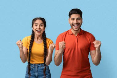 Heyecanlı iki arkadaş mutluluğu ve coşkuyu geniş bir gülümsemeyle ifade eder ve sağlam mavi bir arkaplan önünde dururken yumruklarını kaldırır. Enerjileri neşe verici bir olayı işaret ediyor..