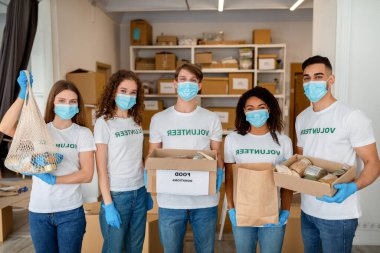 Beş genç gönüllü bir hayır kurumu merkezinde kapalı bir yerde poz veriyor. Eldiven ve maske takarken, yiyecek dolu bağış kutuları tutuyorlar. Takım çalışmaları toplum desteğini destekliyor.
