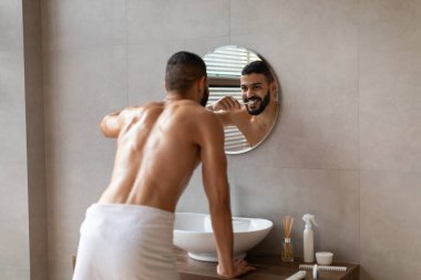 Modern bir banyoda, mutlu genç bir Arap, yuvarlak aynaya bakarken dişlerini fırçalar. Beyaz bir havlu giyiyor ve sağlıklı bir gülümseme sergiliyor..