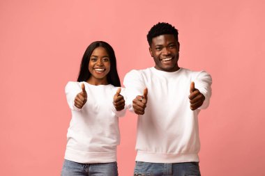 Neşeli bir Afrikalı Amerikalı erkek ve kadın pembe bir arka plana karşı birlikte poz veriyorlar. İkisi de gülümsüyor ve başparmak işareti yapıyor. Birbirine uyumlu kıyafetlerindeki pozitiflik ve mutluluğu gösteriyor..