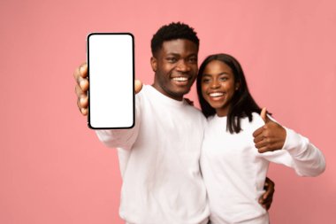 İki neşeli Afro-Amerikan aşığı pembe bir stüdyoda birlikte poz veriyorlar. Boş bir ekran gösteren modern bir akıllı telefon tutuyorlar ve yeni bir mobil uygulama için heyecan öneriyorlar..