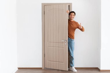 Neşeli genç bir adam evinin kapısında duruyor, gülümsüyor ve baş parmak işareti yapıyor. Ziyaretçileri sıcak karşılayarak evinde dostane bir ortam yaratıyor..