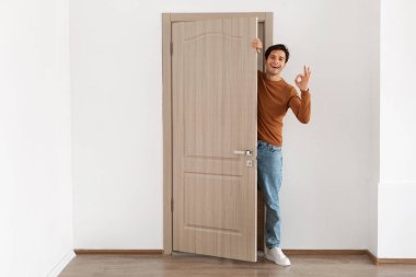 Neşeli genç bir adam evinin kapısında duruyor, gülümsüyor ve dışarı bakıyor. Elleriyle iyi bir işaret veriyor. Sıcak, aydınlık bir mekanda dostça bir tavırla ziyaretçileri karşılıyor..