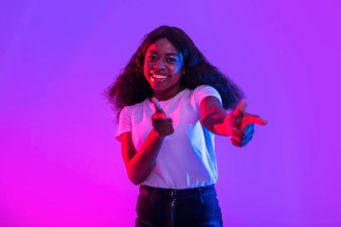 Genç siyah bir kadın parlak neon ışıklarıyla kamerayı işaret ederken parlak bir şekilde gülümsüyor. Mutluluk ve güven saçıyor, seyirciyi neşeli tavırlarıyla meşgul ediyor..