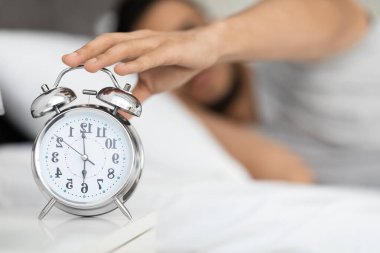 Yatakta uzanmış, komodinin üzerinde klasik bir ikiz çan çalar saatine uzanan bir adamın yakın çekimi. Ayarlar rahat bir yatak odası, sabah rutinini ve uyanışı gösteriyor..