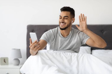 Genç bir Arap, akıllı telefonuyla yatakta oturuyor kulaklık takıyor ve gülümsüyor. Kameraya el sallıyor, arkadaşlarıyla video bağlantısı kuruyor, neşe saçıyor..