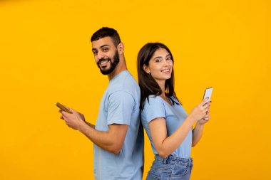 Modern bir Arap çift cep telefonlarını kullanırken sırt sırta verip gülümsüyor. Cihazları ile meşguller, canlı sarı bir stüdyo ortamında çevrimiçi etkileşimlerin tadını çıkarıyorlar..