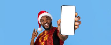 Parlak kırmızı Noel Baba şapkası takan neşeli bir adam iki eliyle boş bir akıllı telefon sergiliyor. Renkli, geleneksel bir kıyafet giymiş ve canlı, mavi bir arka plana karşı neşeli bir şekilde poz veriyor..