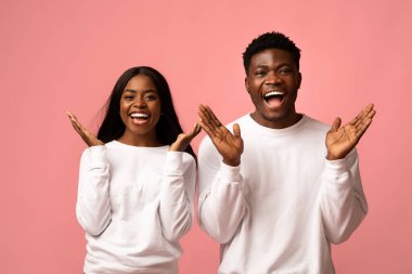 Beyaz tişörtlü genç siyah bir çift ellerini kaldırıp gülümseyerek şaşkınlık ve mutluluk ifade ediyor. Arka plan yumuşak bir pembe, neşeli enerjilerini arttırıyor..