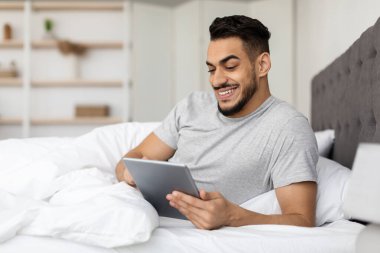 Yakışıklı bir Orta Doğulu genç adam, tabletinde internette gezinirken gülümseyerek yatakta vakit geçiriyor. Konforlu yatak odası ona kendini evinde ve rahat hissettiriyor..