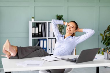 Modern bir ofiste, mutlu bir bayan girişimci, ayakları masanın üzerinde bir koltukta oturarak ara verir. Hayal kurarken gülümsüyor, elleri başının arkasında, kendini rahat hissediyor..