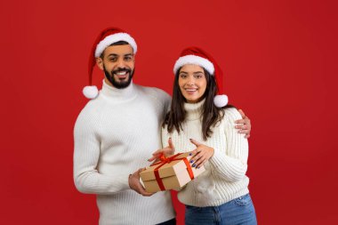 Neşeli Arap eşler bir hediye kutusu tutuyorlar ve kameraya gülümsüyorlar. Noel Baba şapkası takıyorlar. Birlikte Noel 'i kutlamak için harika bir ortam yaratıyorlar..