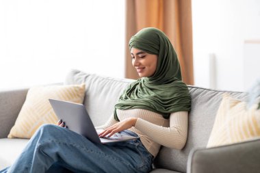 Genç bir Arap kadın tesettüre dizüstü bilgisayar kullanarak rahatça oturuyor. Çeşitli çevrimiçi aktivitelere katılıyor, evde iş ve boş vakitleri dengeliyor..