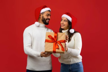 Sevgi dolu bir Arap çift Noel hediyesi kutusunu tutarken birbirlerine gülümsüyor. Her ikisi de Noel Baba şapkası takıyor ve parlak kırmızı bir arka plana rağmen bayram kutlamaları için şenlikli bir ortam yaratıyor..