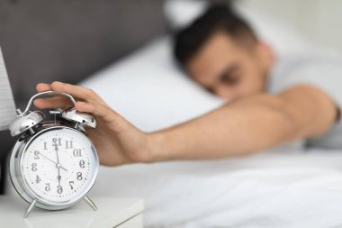 Genç bir Arap adam yatakta uzanıyor, elini komodinin üzerindeki çalar saate doğru uzatıyor. O, sıcak odasında güne başlamak için uykulu ve isteksiz görünüyor. Güneş ışığı sahneyi hafifçe aydınlatır..