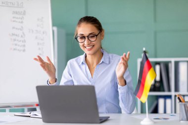 Neşeli bir bayan öğretmen evinden online Almanca dersi veriyor. Sosyal uzaklık pratiği yaparken, interaktif öğrenmeyi teşvik ederken öğrencilerle bir dizüstü bilgisayar üzerinden iletişim kuruyor..