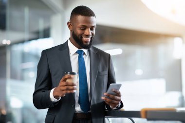 Takım elbiseli bir adam telefonuna bakarken ofiste dikiliyor. Gülümsüyor ve diğer elinde bir kahve fincanı tutuyor. Olay yeri parlak, gündüz olduğunu gösteriyor..