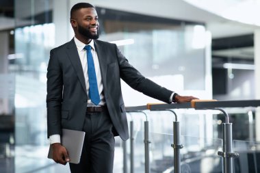 Takım elbiseli profesyonel bir adam ofis binasında duruyor. Bir dizüstü bilgisayar tutuyor ve gülümsüyor. Doğal ışık pencerelerden içeri giriyor ve modern iç tasarımı vurguluyor..