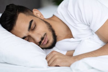 Yakışıklı genç bir Arap, rahat bir yatakta başını yumuşak bir yastığa koyup mışıl mışıl uyuyor. Parlak, beyaz yatak odası gündüz uykusu için sakin bir atmosfer yaratıyor..