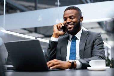Takım elbiseli bir adam telefonuyla konuşurken gülümsüyor ve yoğun bir ofisteki bir masada dizüstü bilgisayar kullanıyor. Bir fincan kahve onun yanındaki masaya konur..