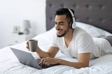 Neşeli bir Orta Doğulu adam kulaklığı açık olarak yatakta dinleniyor ve dizüstü bilgisayarında internette geziniyor. Elinde bir fincan kahve tutuyor ve rahat bir odada dinleniyor..