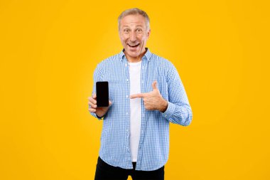Bir adam sarı bir arkaplanın önünde duruyor. Gülümserken elinde tuttuğu akıllı telefonu işaret ediyor. Giysileri sıradan, altında bir tişört ve beyaz tişört var..