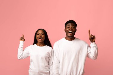 Eşleşen beyaz tişörtlü heyecanlı bir Afrikalı Amerikalı çift işaret parmaklarıyla gülümsüyor ve pembe stüdyo arka planında yaratıcılıklarını ve mutluluklarını ifade ediyorlar..