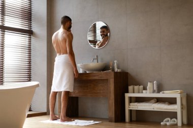 Genç Arap adam modern bir banyoda üstsüz duruyor aynaya bakıyor ve çenesine dokunuyor. Sıcak bir duştan sonra cilt bakımını yansıtan bir havluya sarılmış..