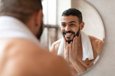 Kendine güvenen genç bir Arap, banyoda aynanın önünde duruyor ve sakalına gülümseyerek dokunuyor. Yansımasından, özgüveninden ve özgüveninden zevk alıyor..