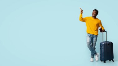 Mutlu Afrikalı Amerikalı genç turist uçak biletleri ve pasaport taşıyor, bavuluyla mavi stüdyonun arka planında poz veriyor, yurtdışına seyahat ediyor, fotokopi alanı olan bir panorama