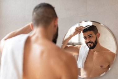 Yakışıklı genç bir adam lüks bir otel banyosunda dikiliyor, yuvarlak aynadaki yansımasına bakarken saçlarını tarıyor. Bakım rutinine odaklanmış görünüyor..