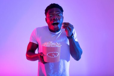 Neşeli genç bir siyah adam renkli, neon ışıklı bir ortamda film izlerken bir kovadan patlamış mısır yiyor. Yüzündeki ifade, film deneyiminin saf heyecanını ve eğlencesini gösteriyor..