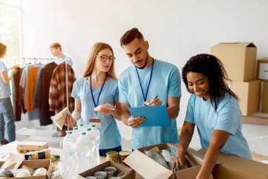 Genç Arap erkek eylemci bağış listesini kontrol ederken diğer gönüllüler yiyecek ve erzak kutularını dolduruyorlar. Sahne, bir hayır kurumu ortamında takım çalışmasını ve toplum desteğini gösteriyor.