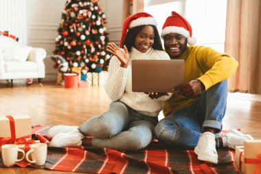 Bir çift yerde dizüstü bilgisayarla oturuyor, gülümsüyor ve video ararken el sallıyorlar. Odada Noel ağacı ve hediyeler var. Bayramlarda şenlikli bir ortam yaratıyor..