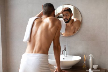 Genç bir Orta Doğulu adam beyaz bir havluya sarılmış bir aynanın önünde duruyor. Saçlarına dokunurken gülümsüyor, iyi aydınlatılmış banyo ortamında yansımasının tadını çıkarıyor..