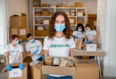 Kadın gönüllü, erzak dolu bir bağış kutusunu tutarken gülümsüyor. İş arkadaşları arka planda daha fazla bağış yapıyor, hepsi güvenlik için maske takıyor..